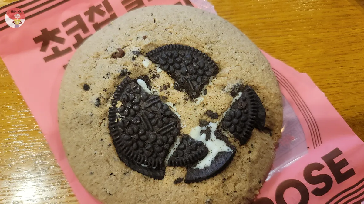 핑크색 포장지 위에 올려진 초코칩 블랙 & 화이트 쿠키, 위에는 부서진 오레오 조각들이 X 모양으로 배열되어 있음