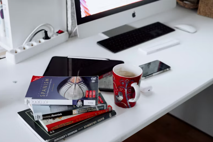 흐트러진 책상 위에 책, 노트 등이 어지럽게 놓여 있고 옆에 스마트폰과 태블릿, 커피잔 등이 놓여 있는 장면