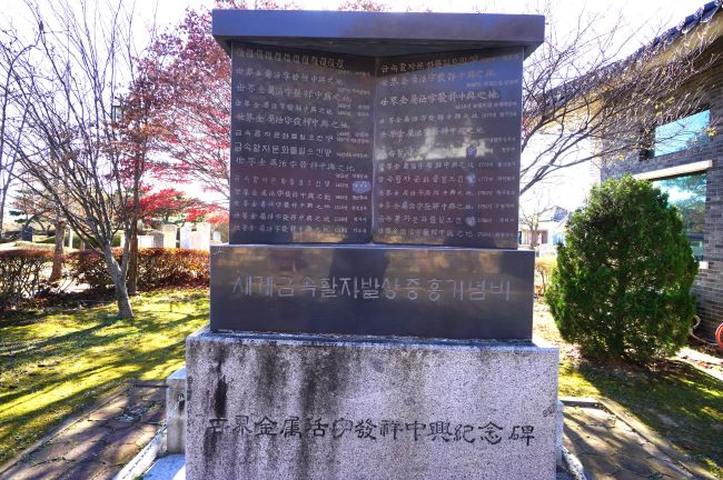 세계금속활자발간중흥기념비, 짙은 회색 대리석에 흰글씨, 아래는 흰색 대리석에 검은 글씨(한자), 뒤로는 공원 풀밭에 나무 그림자들,