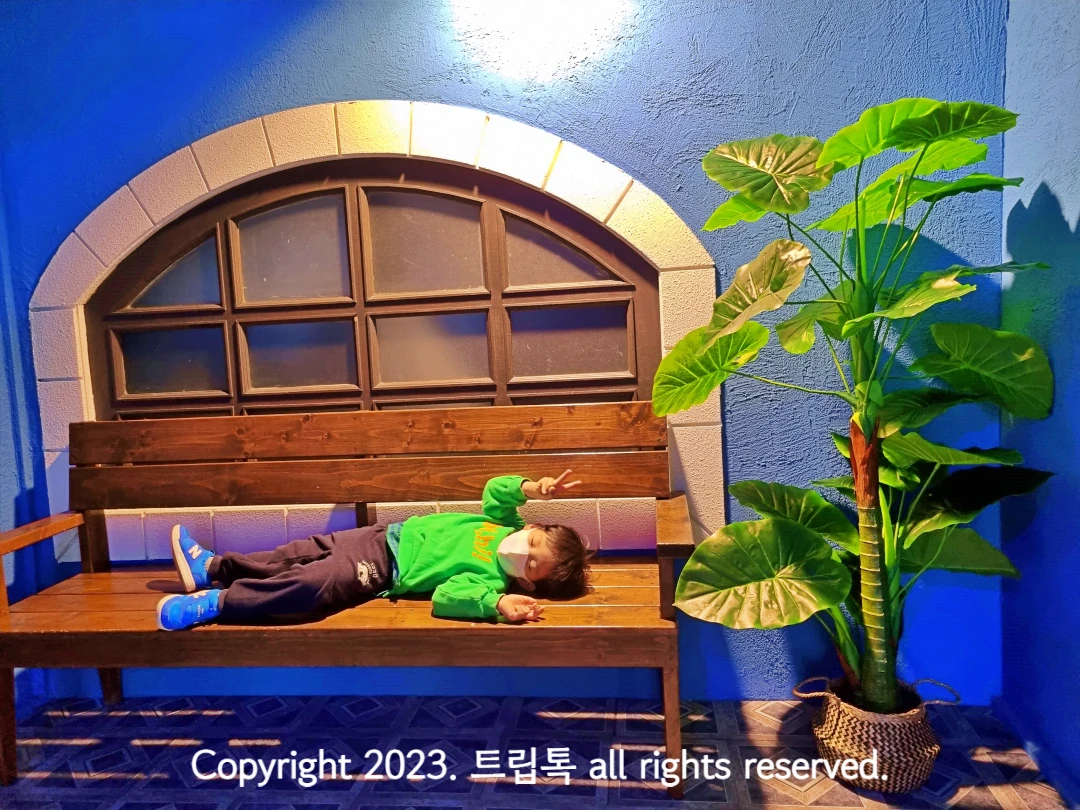 파란 벽에 둥근 창문이 있고 그 아래 벤치에 아이가 누워있다.