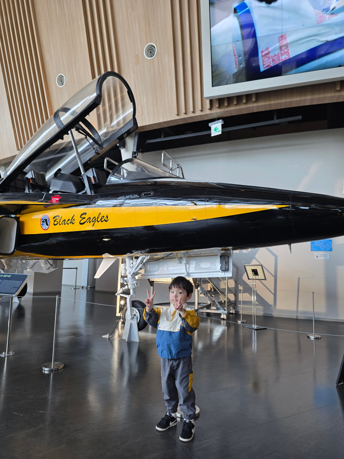 국립항공박물관 전시관에서 블랙이글스 전투기 앞에 서서 브이 포즈를 취하는 어린이. 실제 항공기 모형 앞에서 기념 촬영 중인 모습.