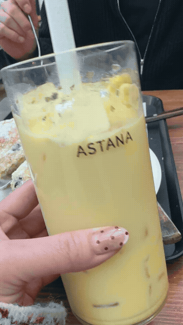 동탄 카페 아스타나에서 주문한 아이스 옥수수라떼를 손으로 들어 올린 장면, 노란빛의 옥수수라떼 위에 거품이 살짝 올라가 있고 컵에는 ASTANA 로고가 보이며 빨대로 저어 마시는 모습이 담긴 GIF