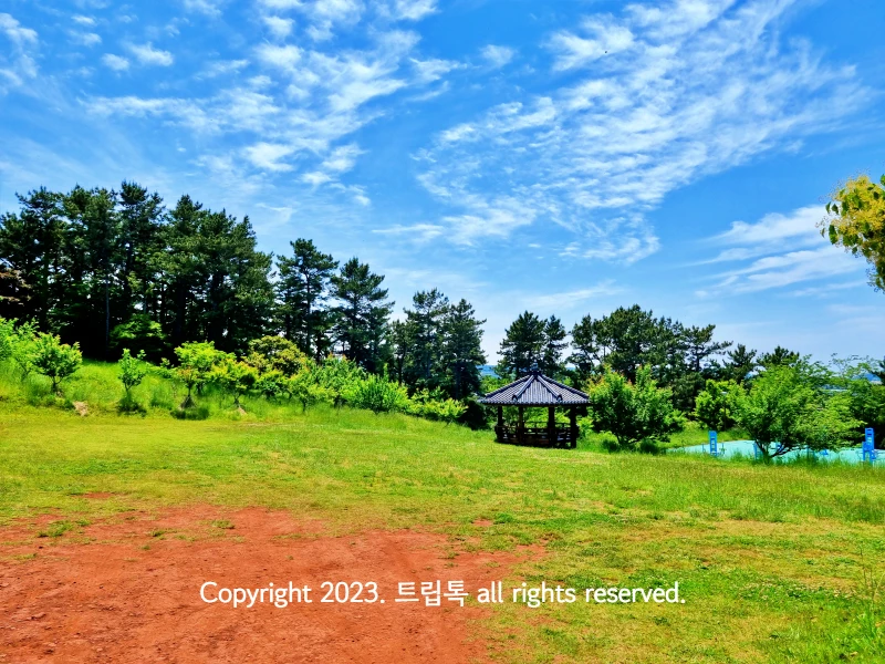 파란 하늘과 하얀 구름아래 정자가 있다. 연두색 풀밭과 붉은 흙이 보인다.