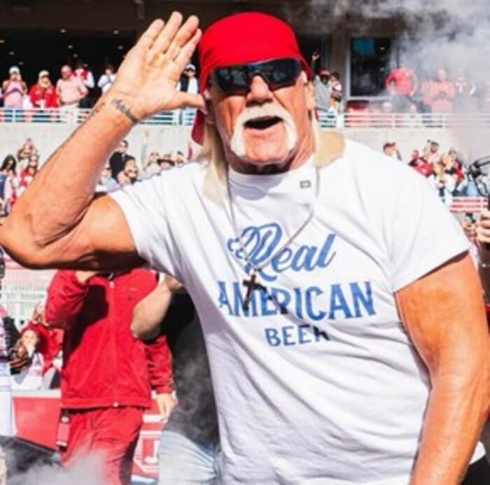 헐크 호건이 야외 경기장 관중 앞에서 오른손을 귀에 대며 팬들의 환호를 유도하는 모습. 흰색 티셔츠와 빨간 반다나, 선글라스를 착용했다.