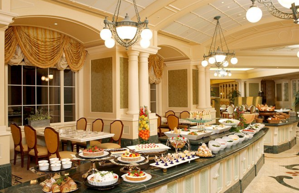 다양한 음식들이 뷔페에 전시되어 있고 뒤로는 테이블들이 보인다.