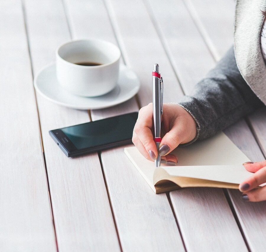 흰색 나무 테이블 위에서 한 여성이 펜을 들고 작은 노트에 글을 쓰고 있는 모습. 옆에는 하얀색 커피 잔과 검은색 스마트폰이 놓여 있음.