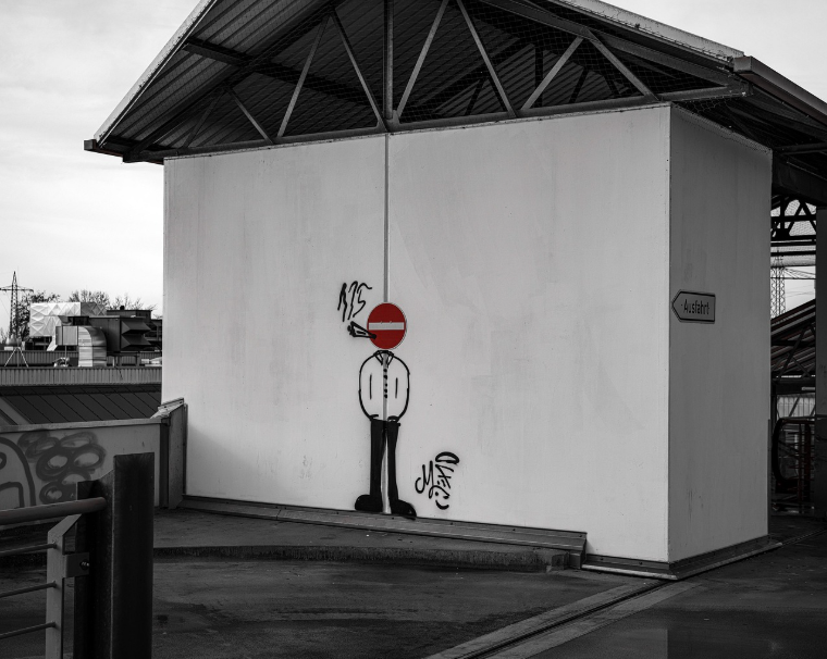 흰 벽면 위에 그려진 거리 예술로, 빨간색 진입 금지 표지판을 머리로 한 인물이 입에 담배를 물고 연기를 내뿜는 모습. 흡연의 위험성과 금지를 풍자적으로 표현한 금연 캠페인 이미지.