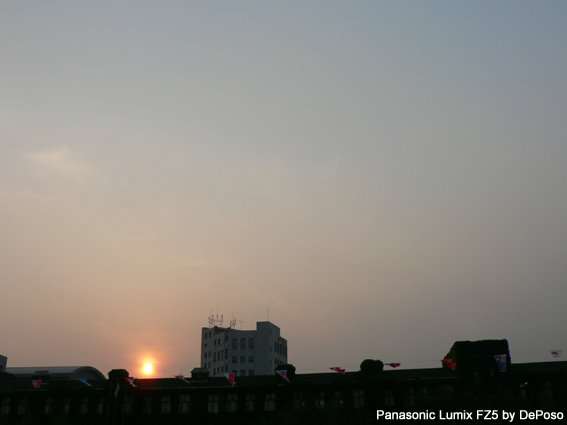 석양이 지는 도시 풍경. 어슴푸레한 하늘 아래, 오래된 건물의 지붕 위에는 여러 나라의 국기들이 장식되어 있고, 그 뒤편으로 현대적인 흰색 건물이 보인다.