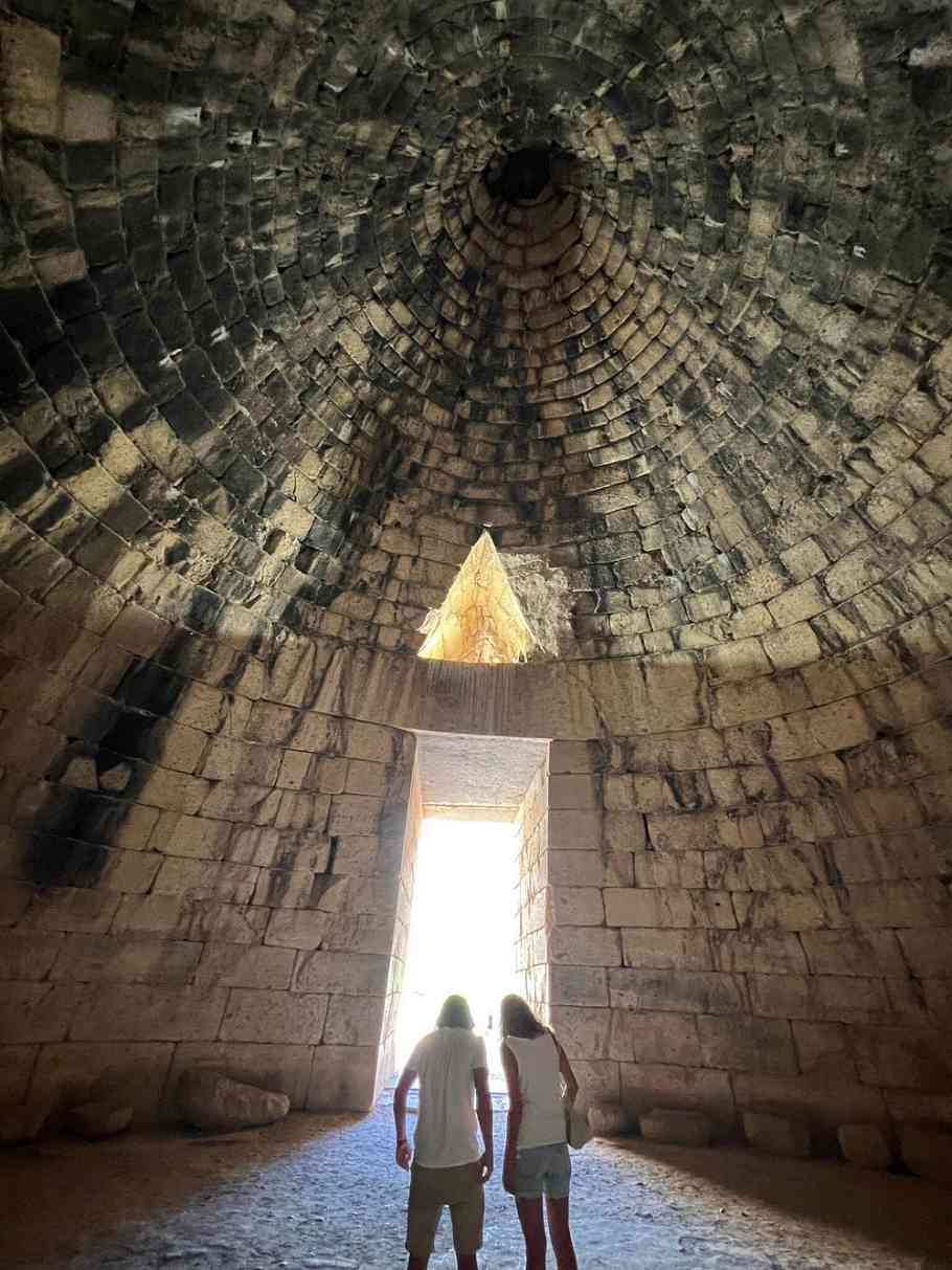 아트레우스의 보고 내부에서 입구를 바라본 장면. 원뿔형 돌 천장과 삼각형 창을 통해 빛이 들어오고, 두 명의 방문객이 출입구 근처에 서 있다.