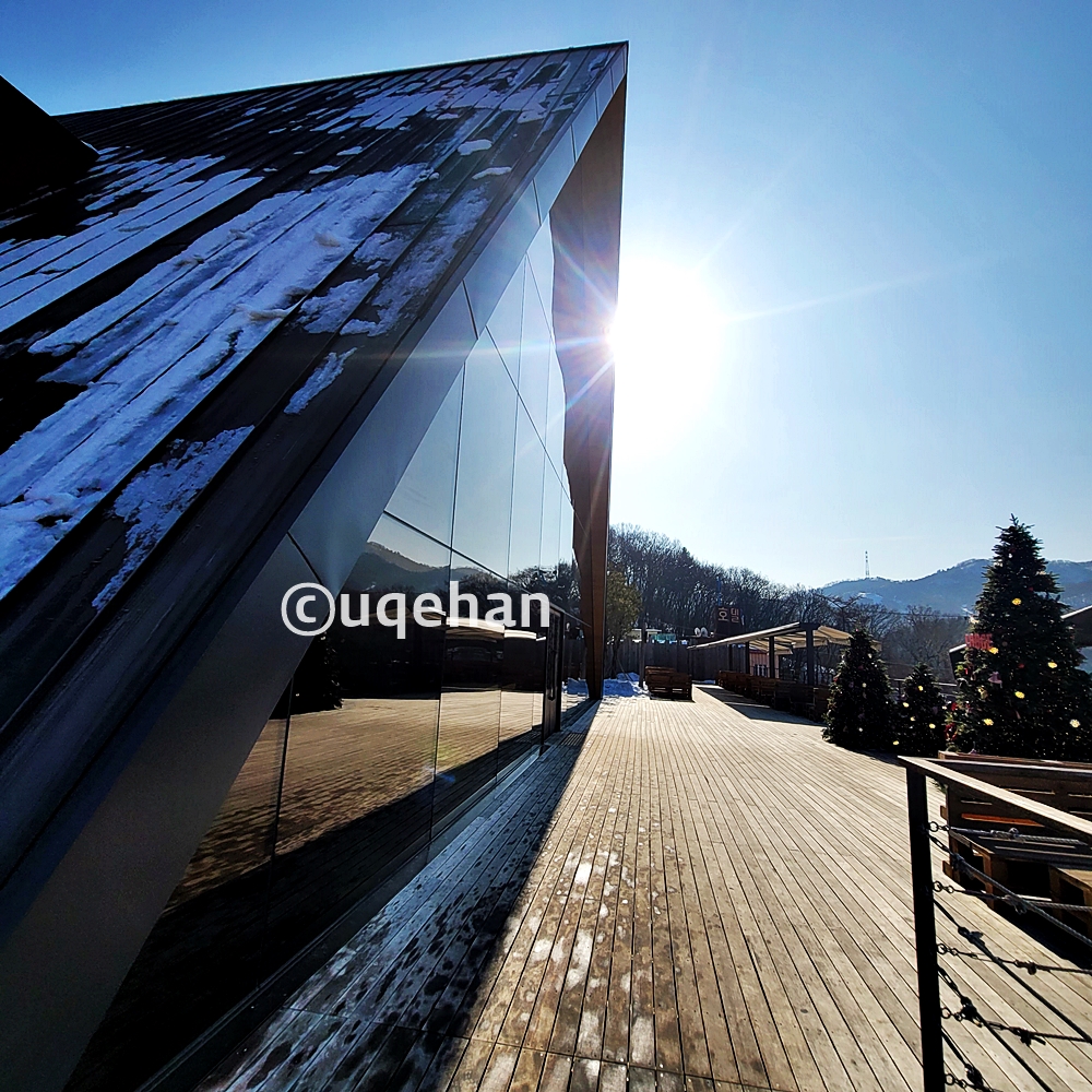 데크위에 건물에 삼각형 모양의 건물이 있다. 오른쪽으로는 야외 테라스 좌석들이 놓여져 있따. 트리도 세워져있다.