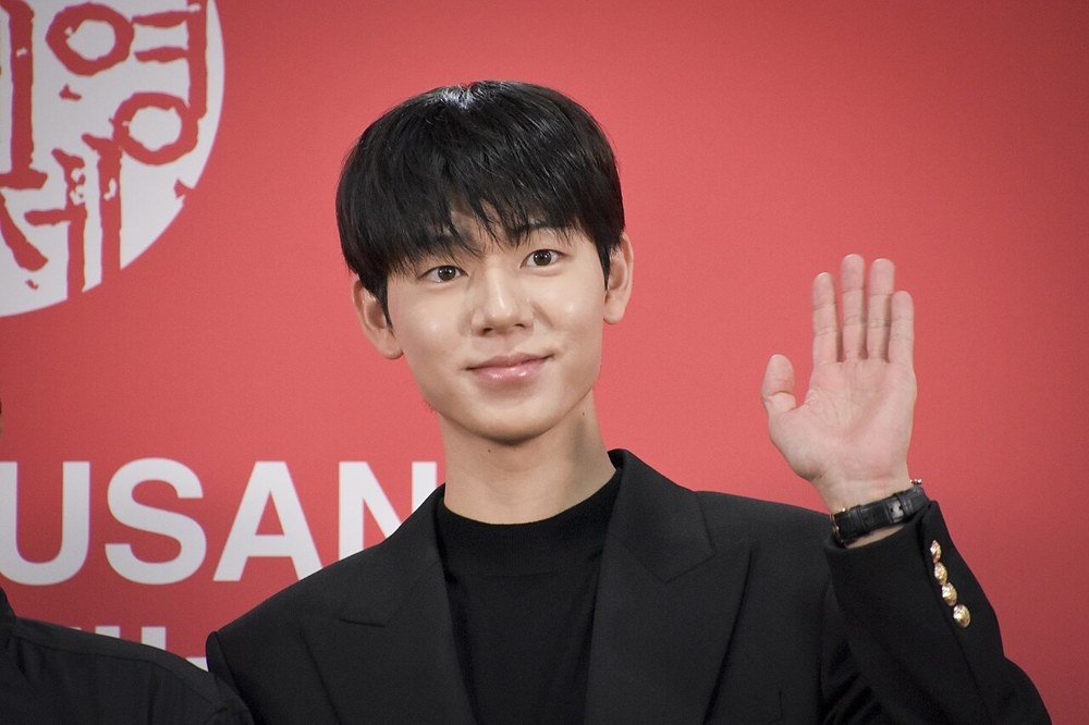BUSAN 글자가 적힌 붉은 배경 앞에서 검은 재킷을 입은 배현성이 한 손을 들어 인사하는 모습
