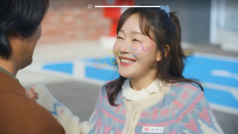 밝은 야외 공간에서 마광숙(엄지원)이 활짝 웃고 있다. 그녀는 흰색 블라우스 위에 부드러운 니트 스웨터를 입고 있으며&amp;#44; 파란색 머리끈으로 머리를 묶었다. 그녀 앞에 있는 남성의 뒷모습이 살짝 보이며&amp;#44; 두 사람이 대화를 나누는 듯하다.