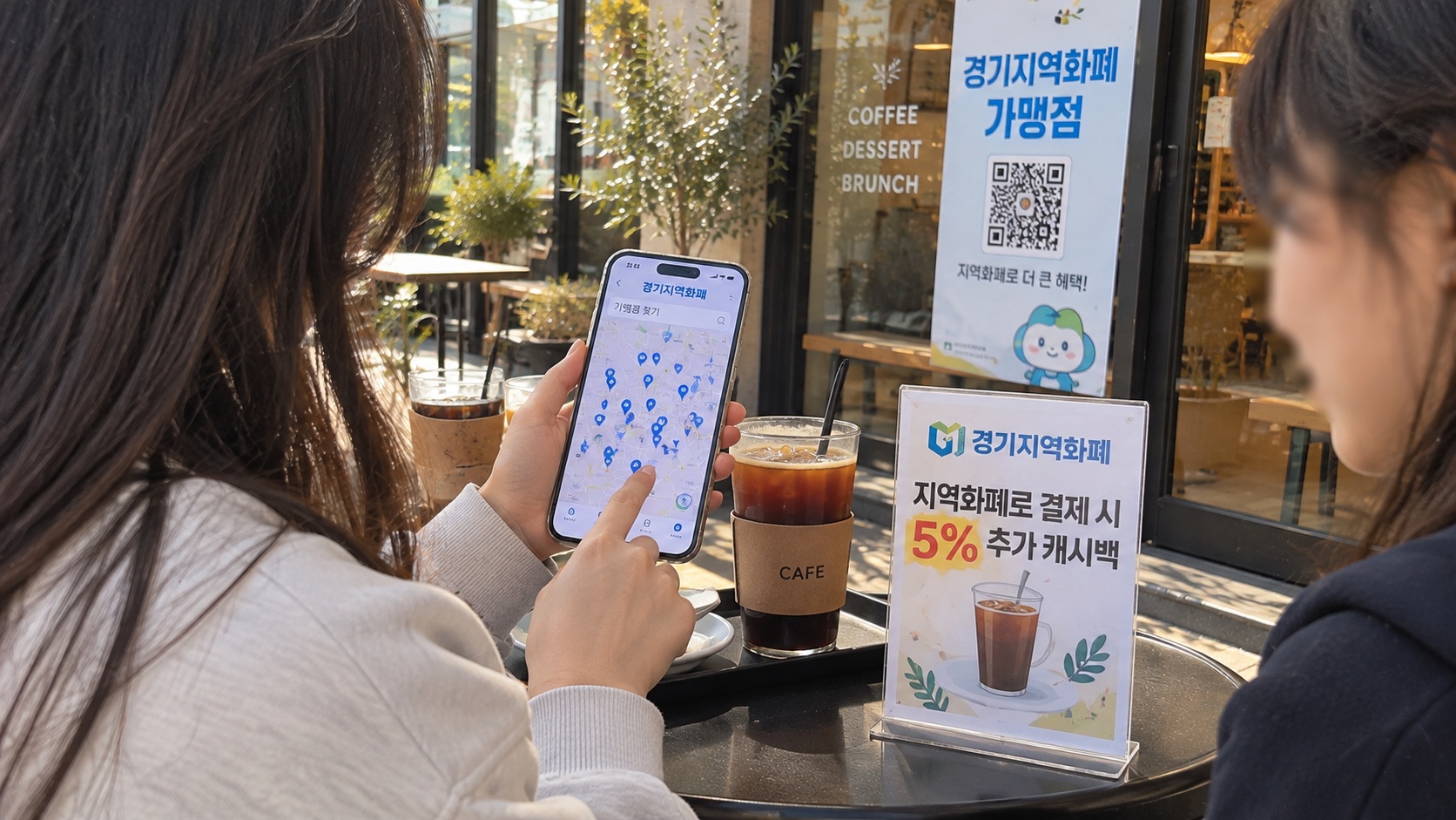 카페 테라스에서 스마트폰 경기지역화폐 앱의 가맹점 찾기 지도를 확인하는 모습과 지역화폐 캐시백 안내판