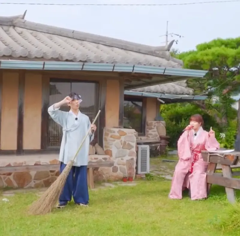 악뮤-전통가옥 앞 잔디위 좌측 파란색 머슴옷에 싸리비를 들고 있는 찬혁과 오른쪽 분홍색 옷을 입고 노래하고 있는 수현