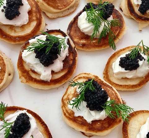 노릇하게 구워진 작은 팬케이크 10개 위에 크림버터와 검정색 캐비아들이 올려졌고, 그 위에는 초록색 로즈마리가 있다.