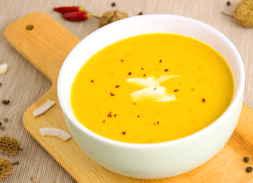 나무 쟁반위에 하얀 죽그릇이 있다. 그릇에는 노란 호박죽이 있고 주변에는 말린 빨간 고추 2개가 있다.