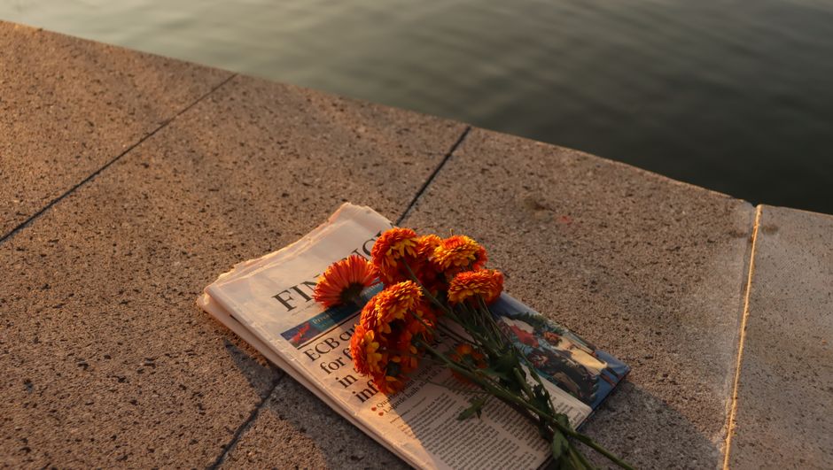 A serene scene of orange flowers placed on a newspaper by a waterside at sunset.
