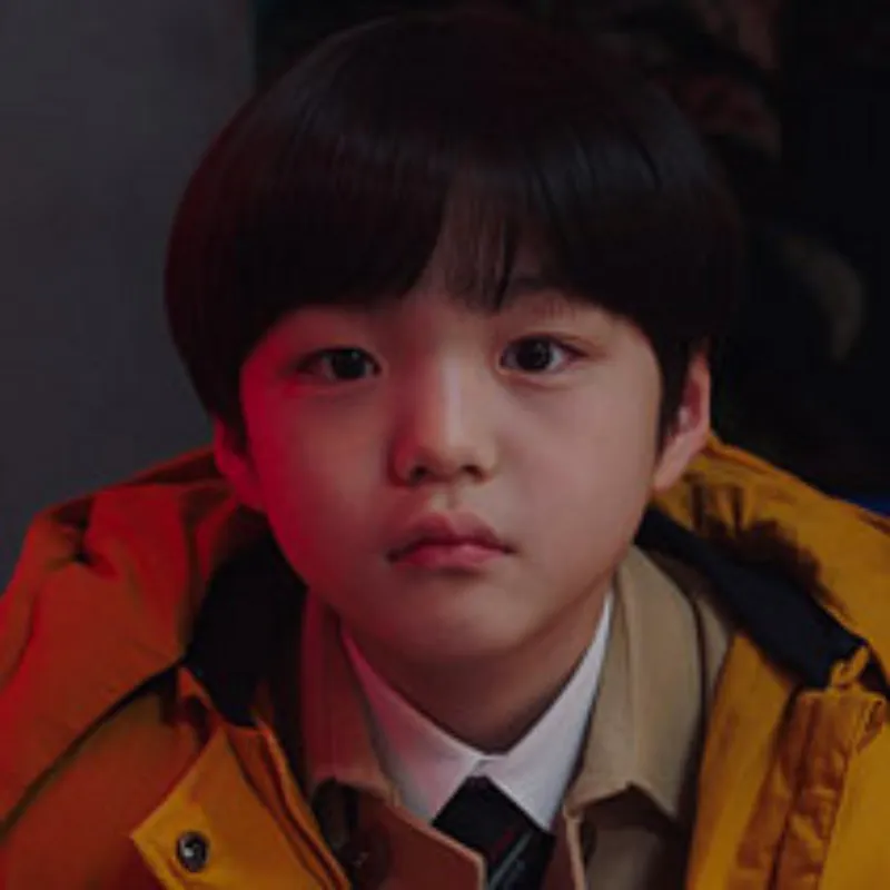 어느 실내를 배경으로 노란 겨울 점퍼를 입고 누군가를 빤히 보고 있는 어린 소년 김민재