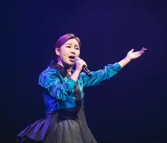 송가인- 무대위 검은 치마 파란 저고리 오른손으로 마이크를 들고 왼손은 하늘을 가리키며 노래하는 송가인