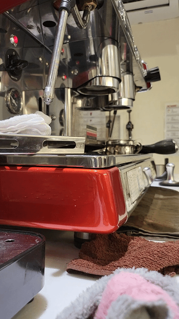 카페 마감 후 커피 머신 그룹헤드에 세척 약품을 넣고 '자동 세척(백플러싱)' 기능이 작동하며 흰 거품이 뿜어져 나오는 역동적인 GIF 이미지. 사장이 직접 현장에서 밤늦게까지 장비를 닦아내는 '지옥의 숙련기'를 시각적으로 증명함.