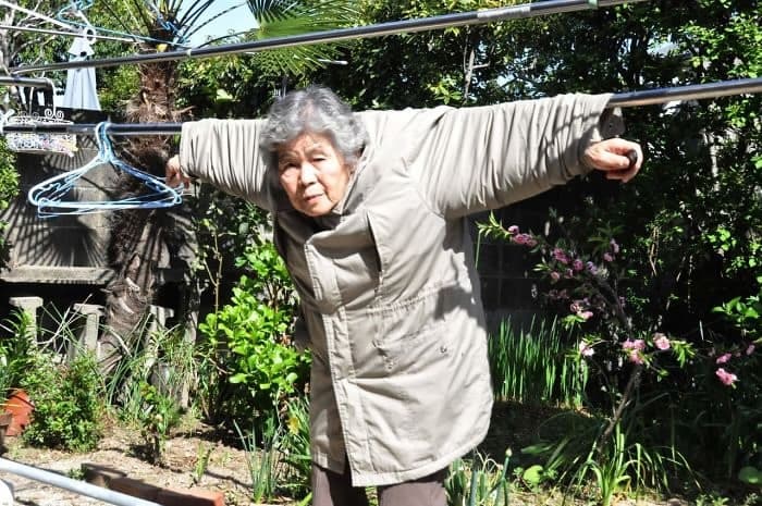허벅다리까지 오는 베이지 색의 잠바를 입고 푸른 나무와 붉은 꽃이 피어있는 정원 앞에 걸린 빨랫줄에 자신의 양팔을 벌린 채, 옷 사이로 빨랫줄을 넣어 자신을 빨랫줄에 걸어 놓은 할머니 모습 이미지