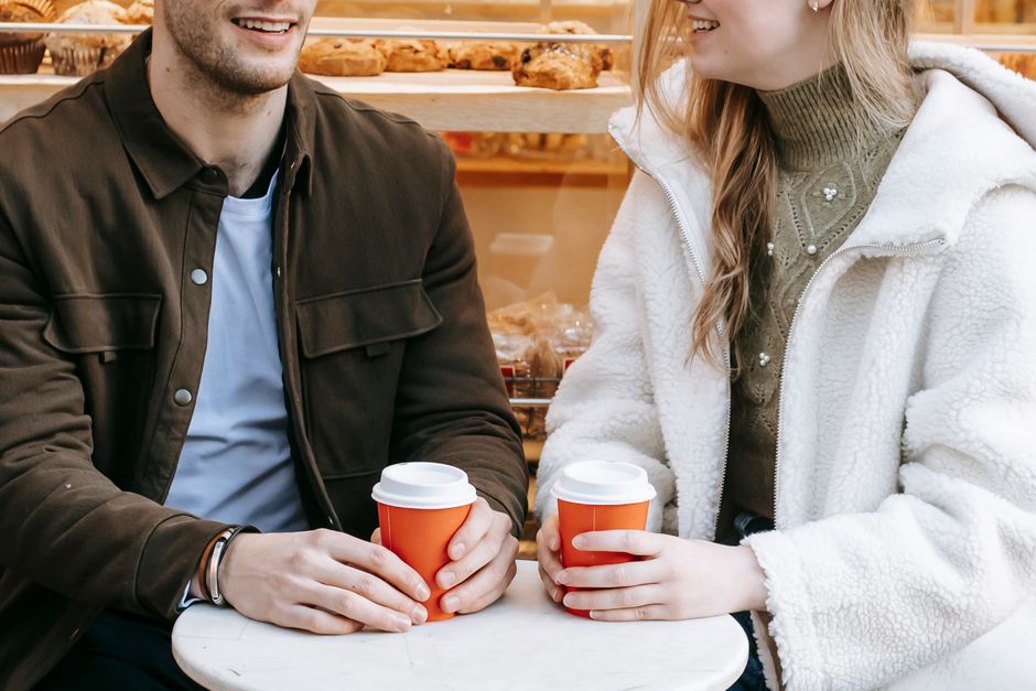 Unrecognizable happy couple in casual wear sitting at small table and having hot beverage in paper cup in cafe