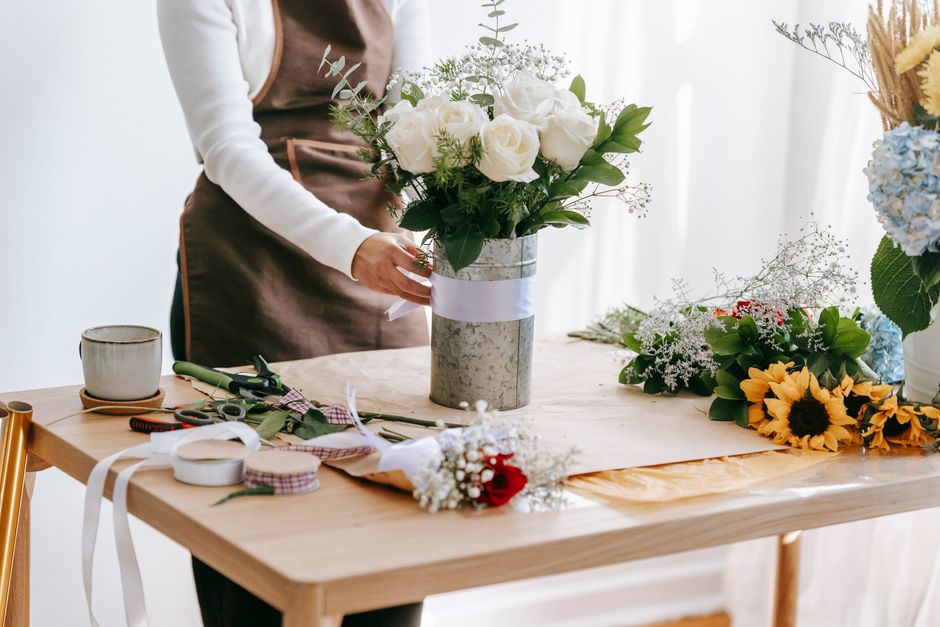 Crop anonymous female florist in brown apron standing and tying ribbon on vase with elegant bouquet with white roses in light room in daytime