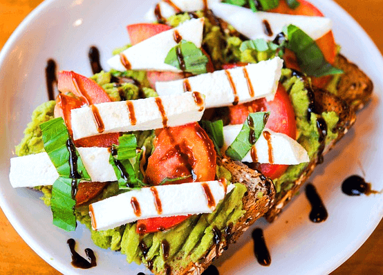 하얀 접시에 샐러드와 치즈를 얹은 식빵이 있다. 빵 위에 아보카도, 토마토, 하얀 치즈가 있고 그 위에는 고동색의 발사믹식초가 뿌려졌다.