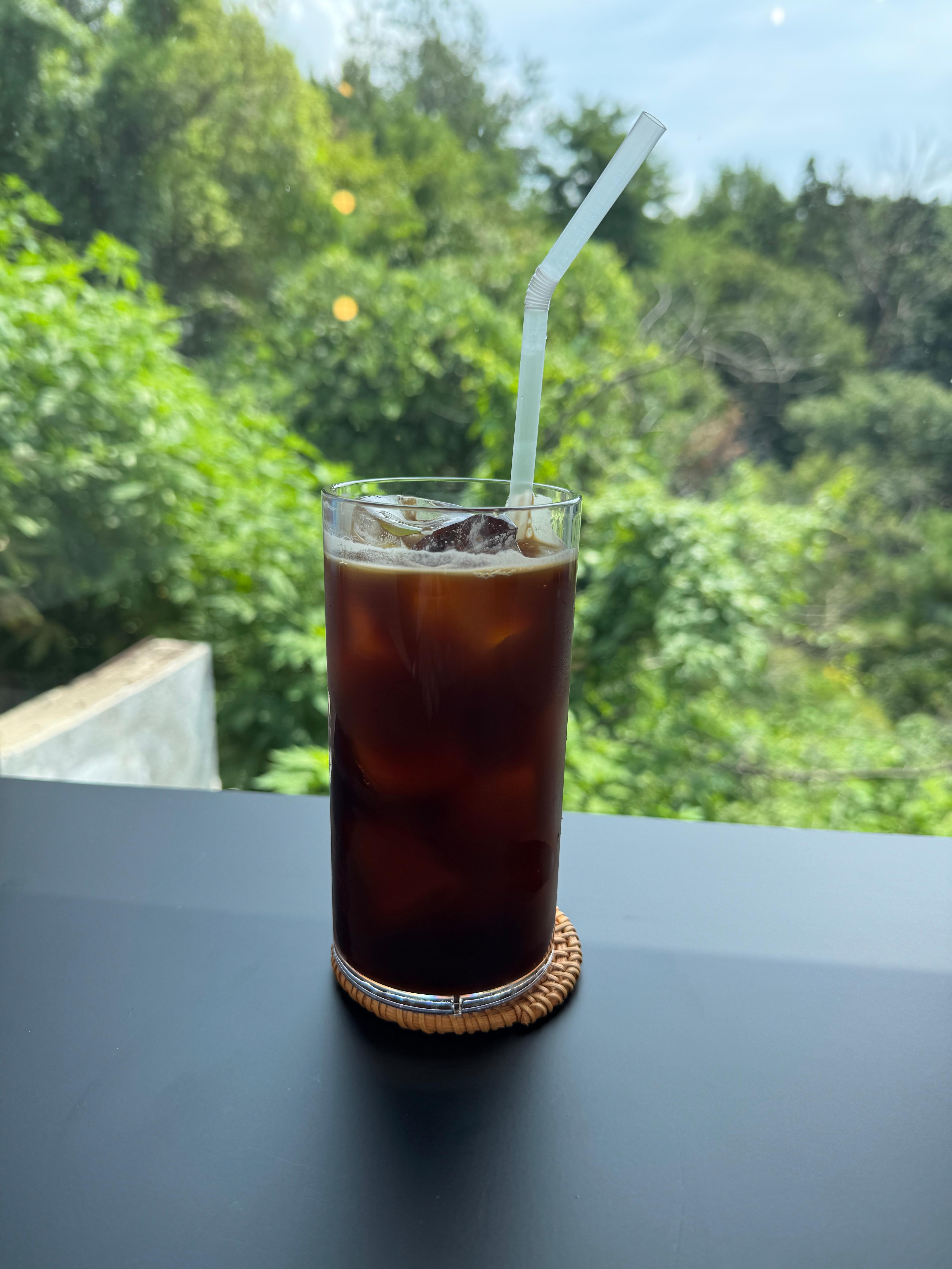 나무가 보이는 창밖을 배경으로 아이스아메리카노 사진을 촬영함