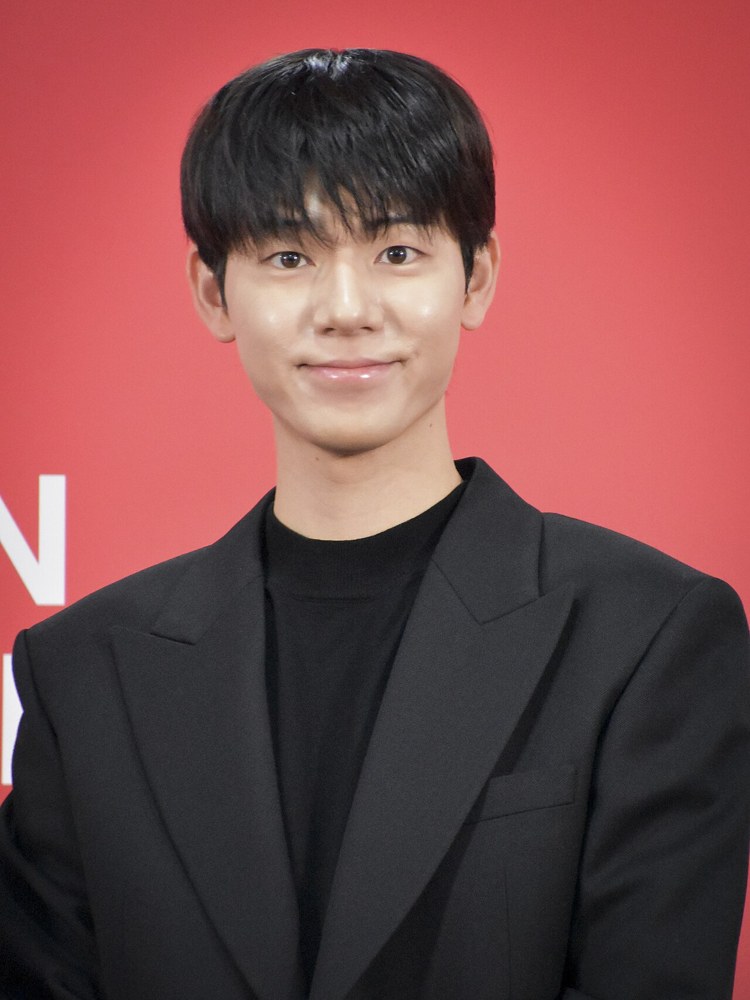 붉은 배경 앞에서 검은 재킷과 검은 상의를 입고 미소 짓는 배현성의 상반신