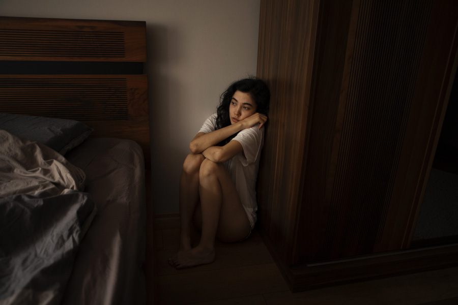 서양의 젊은 여성이 심신불안증으로 침대 옆의 구석에 쪼그리고 앉아서 한 곳을 응시하며 불안해 하고 있는 것을 찍은 사진