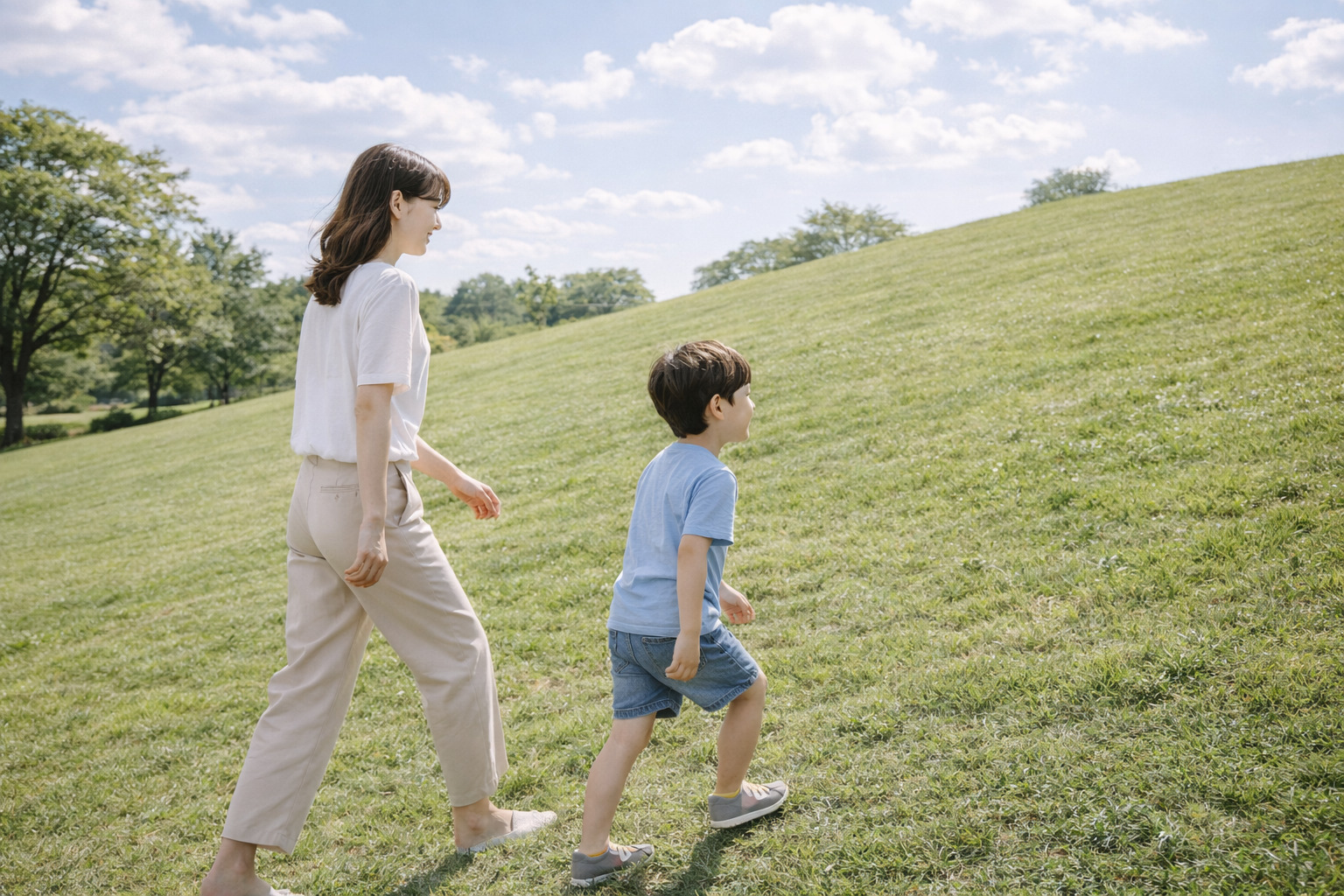 초록 잔디 언덕에서 엄마의 뒤를 따라 걷는 6살 남자아이의 옆모습이 담긴 평온한 장면