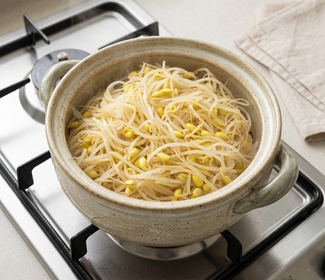 주방 가스레인지 위에서 김이 모락모락 피어오르는 오덴세 풍의 베이지색 무광 도자기 솥. 솥 안에는 하얀 밥 위로 노란 머리의 콩나물이 소복하게 쌓여 뜸이 들고 있는 요리 과정 사진.