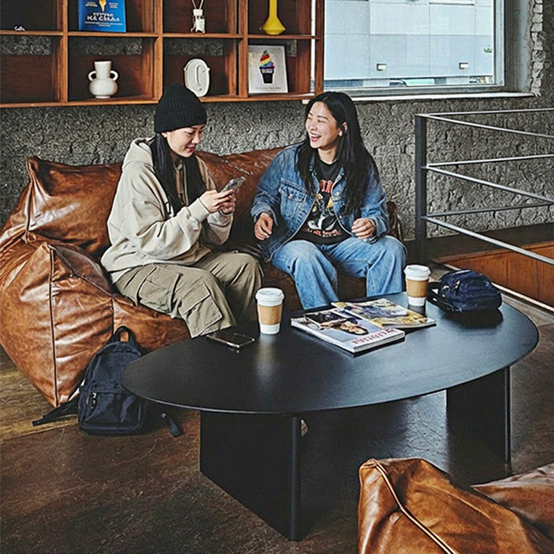 책장과 원형 테이블이 있는 공간에서 두 사람이 플래지어 톰빈백 2인용 갈색 기능성 가죽 빈백에 나란히 앉아 웃으며 이야기하는 모습