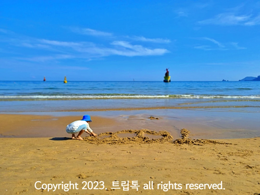 푸른 하늘 아래 바다가 있고 그 아래 모래사장에서 파란 모자를 쓴 아이가 모래놀이 하고 있다