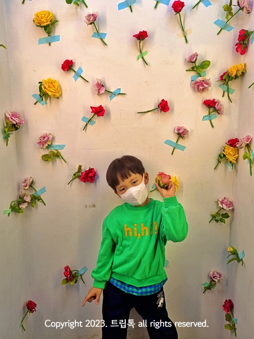 꽃송이들이 하나씩 테이프로 벽에 붙어있고 그 아래 아이가 꽃을 들고 있다.
