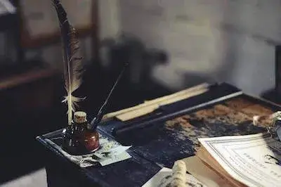 글씨가 인쇄된 종이들이 놓여있는 잉크가 묻어있는 오래된 나무 책상 위 모서리에 놓여있는 잉크병과 병에 꽂혀있는 펜과 깃털이 달린 펜.