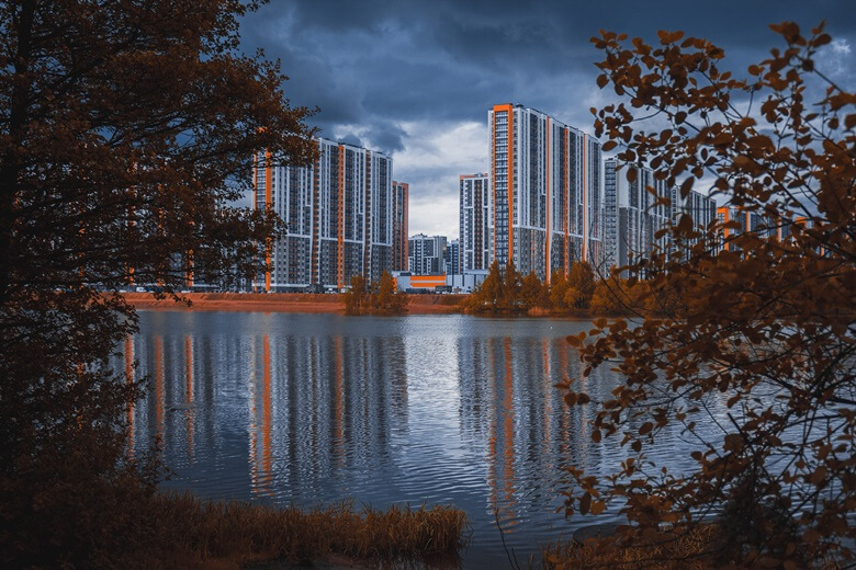 좌/우측으로 갈색으로 물든 나무가지 중앙으로 아래는 강물이 잔잔하게 흐르고 그 뒤로 오랜지색 띠를 두른 하얀색 아파트 단지가 보이고 강물위에 아파트 단지가 거울처럼 비치는 사진