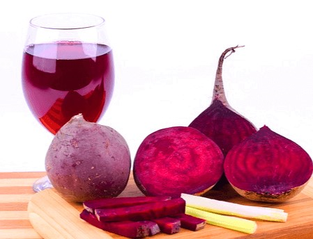 나무 도마 위에 동그랗고 보라빛이 도는 붉은색의 비트 한 덩이와 반으로 잘린 비트 3덩이가 있다. 뒤에는 유리잔에 자주색의 비트주스가 가득 담겨있다.
