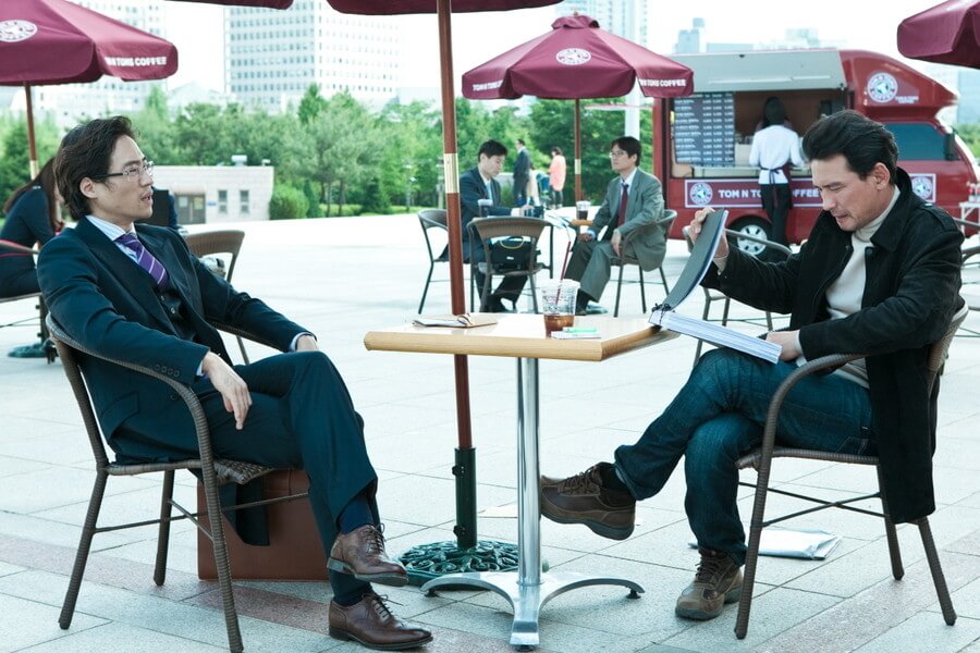 야외 테이블에 앉아 서류를 보고 있는 최철기와 그를 바라보는 주양