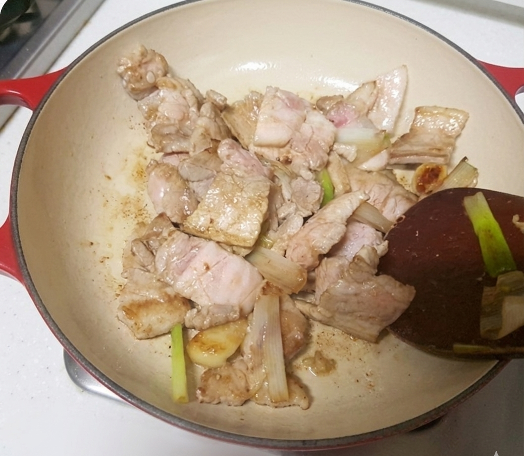 무쇠 팬 위에서 대파와 함께 노릇하게 볶아지고 있는 삼겹살의 근접 촬영 사진. 나무 뒤집개를 사용하여 돼지고기를 볶는 조리 과정 모습.