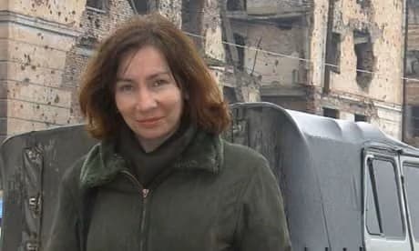 나탈리아 에스테미로바. 한쪽에 차량 한 대가 주차 되어 있는 건물들 사이, 거리를 지나가는 카메라 쪽을 응시하며 의미를알 수 없는 살짝 미소 지은 표정의 이미지.