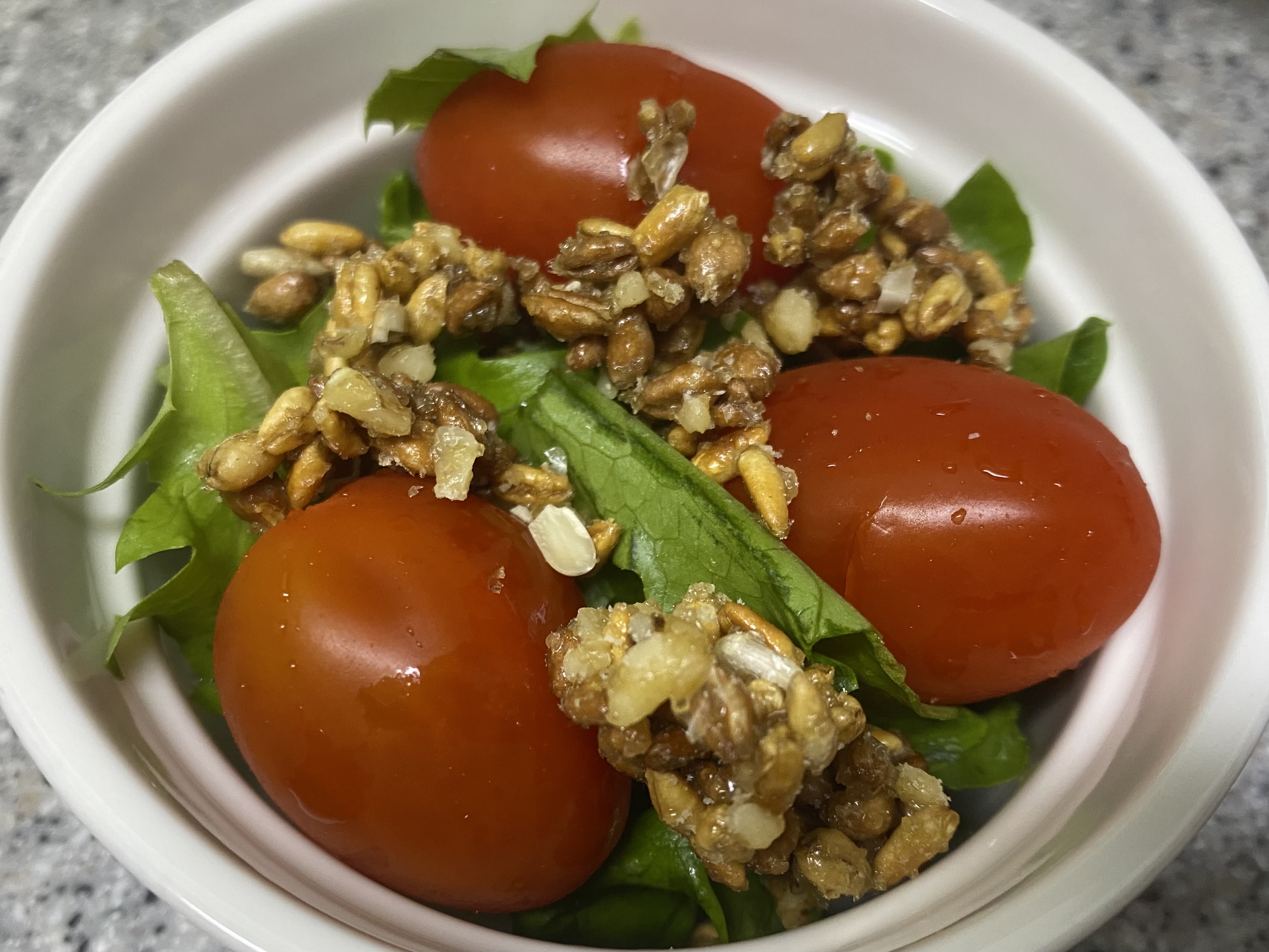 흰색 동그란 그릇 안에 샐러드 야채와 토마토 3개가 있고 '그래놀라 파로이츠'를 토핑한 사진입니다.