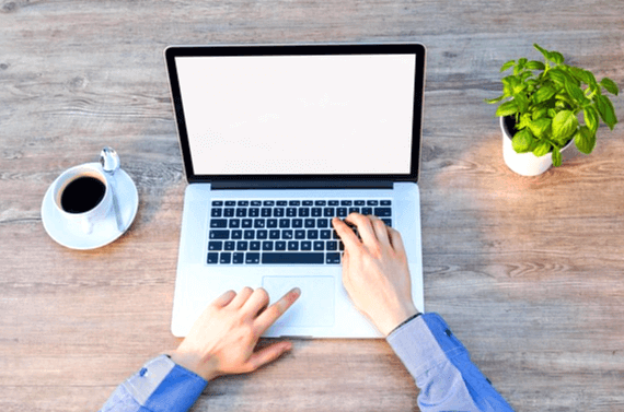 나무 책상에 노트북이 있다. 파란색 셔츠를 입은 남자 손이 키보드를 치고 있고, 블랙커피와 녹색식물 화분이 양쪽에 있다.