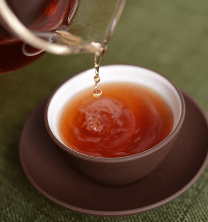 갈색 도기 그릇에 연갈색의 보이차가 가득하고 투명 유리 주전자에서 보이차가 찻잔에 떨어지고 있다.