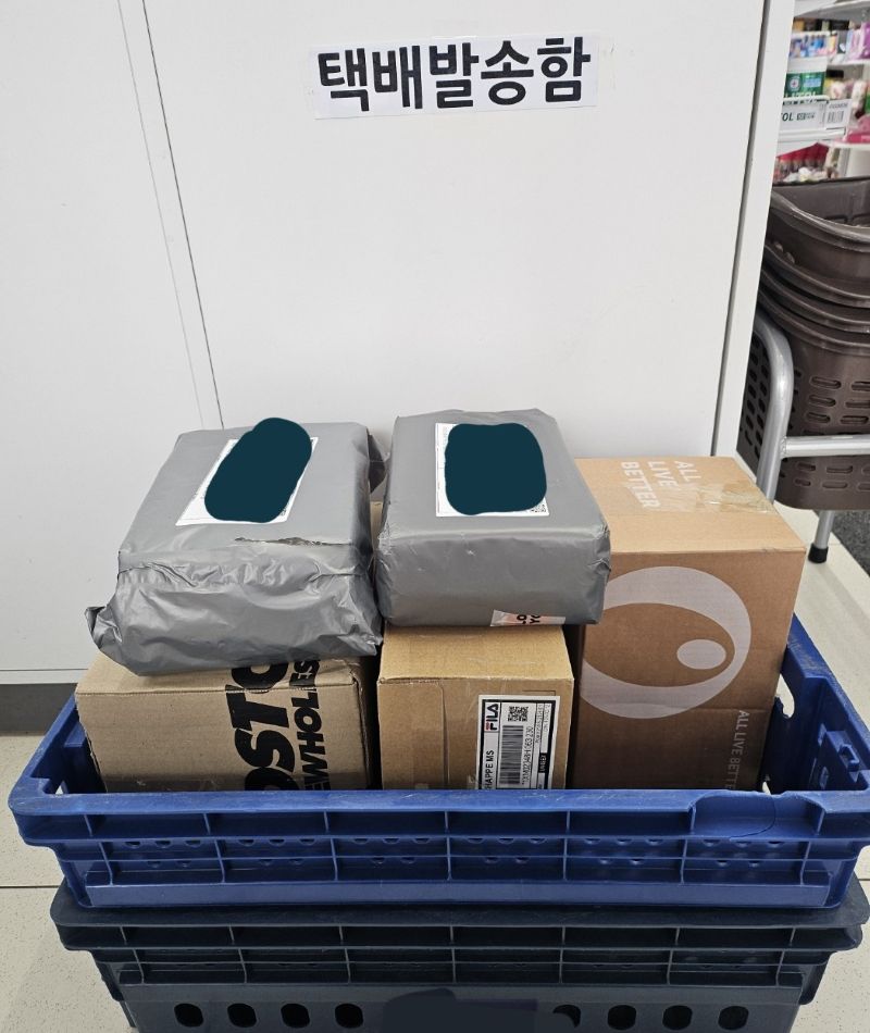 편의점 매장 한구석, 파란색 물류 바구니 위에 수거를 기다리는 다양한 크기와 형태의 택배 박스들이 쌓여 있다. 뒤편 흰색 벽에는 '택배발송함'이라는 안내판이 붙어 있다.