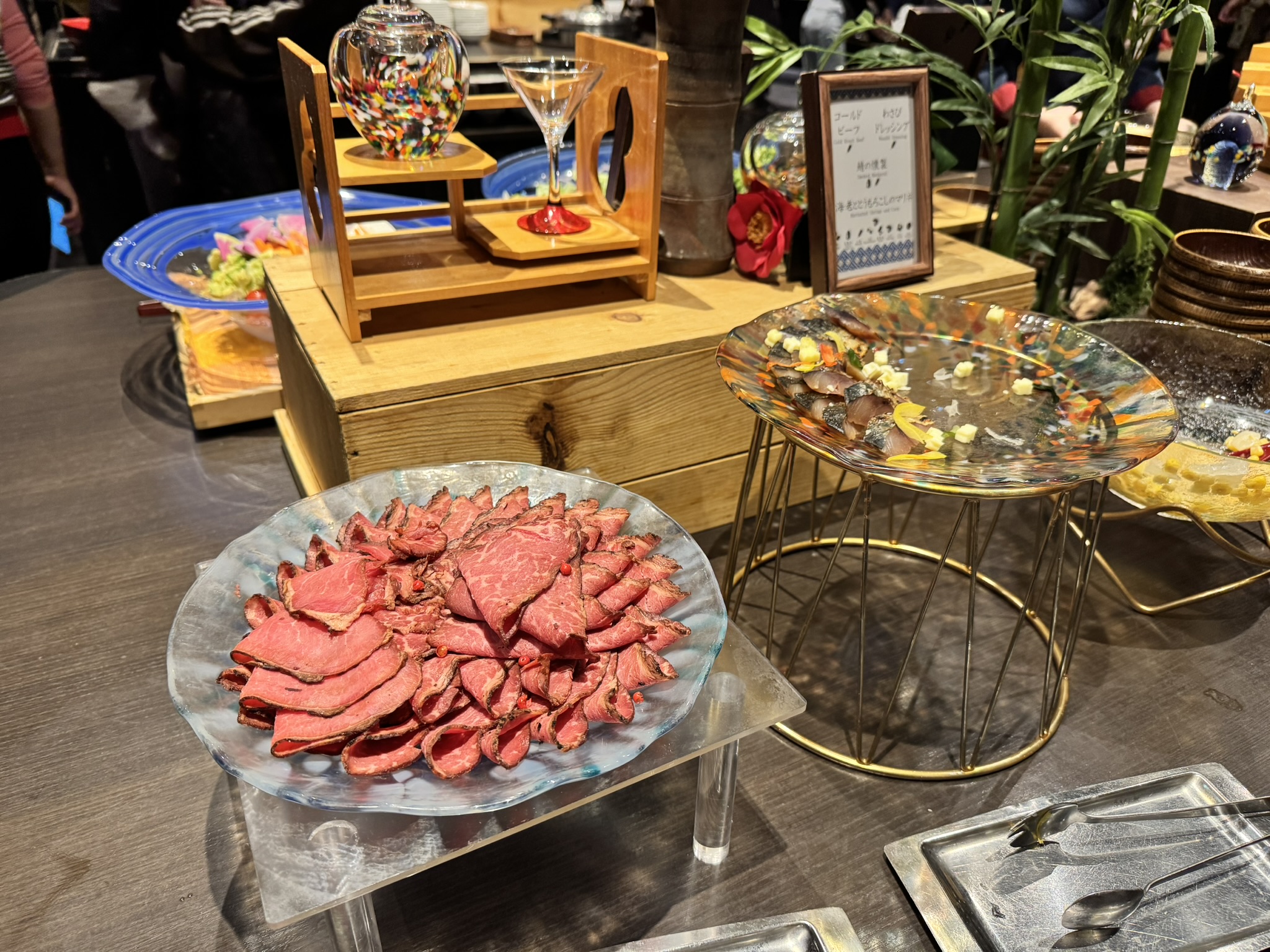 뷔페 테이블에 놓인 얇게 썬 로스트 비프가 가득 담긴 유리 접시와 고등어 초절임(시메사바) 요리. 뒤편으로 화려한 유리 공예 소품과 대나무 장식이 보임.