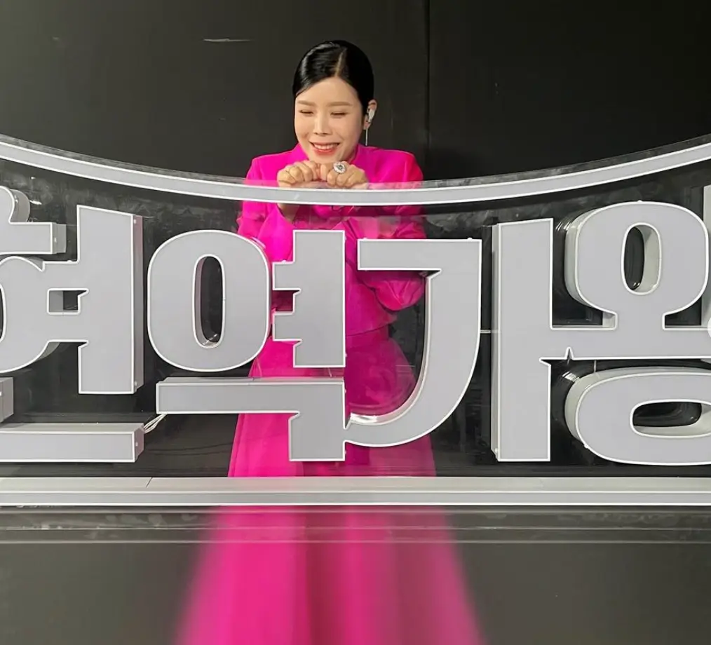 한일가왕전-흰색 한일가왕전글자 판 뒤에서 글자판에 기대어 미소지슨 린