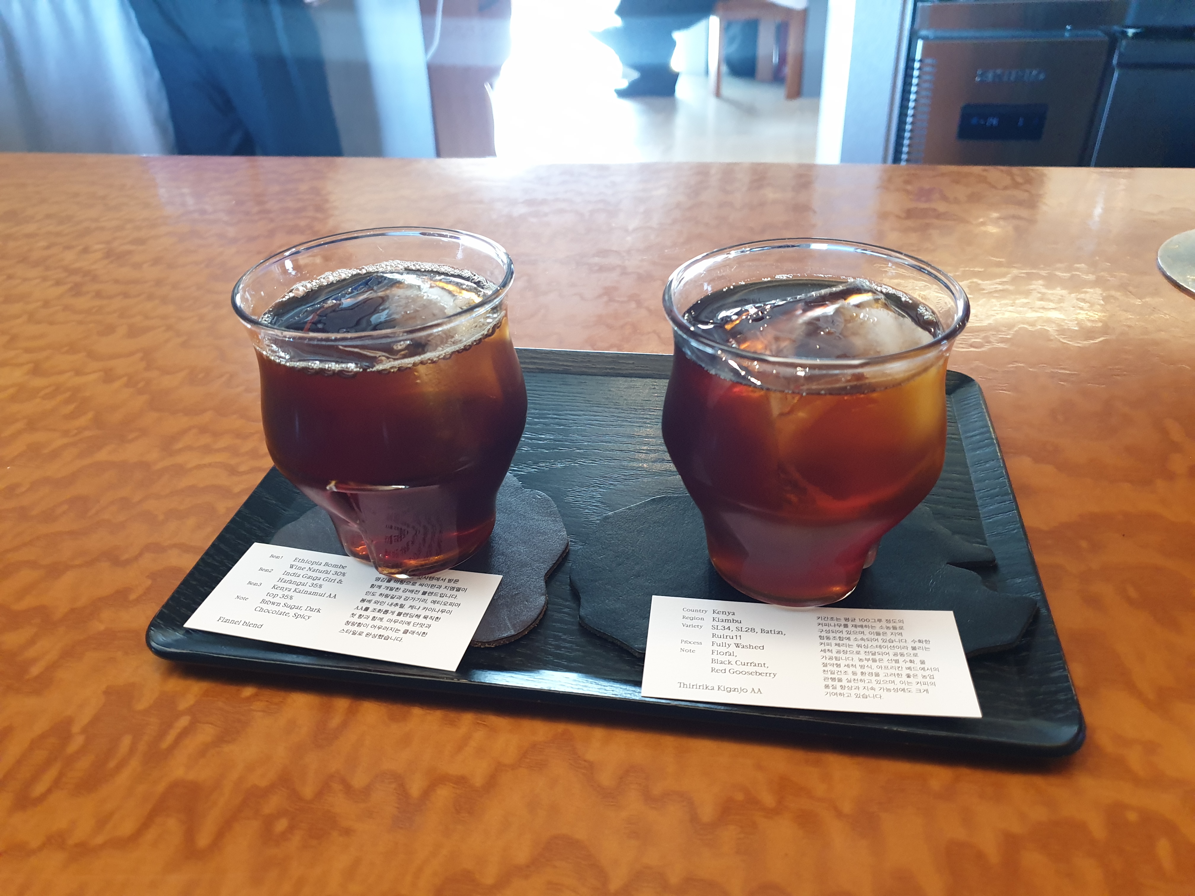 우드 테이블에 검은 쟁반이 놓여있다. 그 쟁반위에 컵 두개에 커피가 담겨있다. 각 커피에는 원두 설명 카드가 놓여있다.