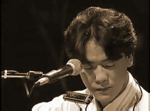 김광석이-목에-하모니카를-메고-마이크-앞에-앉아-눈을-지그시-감고-기타를-연주하는-사진